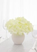 A pack of 50 pale yellow 6.69'' Hydrangea Flower Heads With Stems is arranged in a white vase on a table with a white cloth, surrounded by empty wine glasses in a bright, softly-lit setting.
