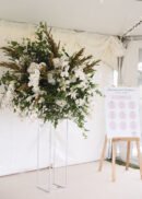 A large floral arrangement with white flowers and greenery sits atop a 32" tall gold metal wedding flower stand with an acrylic board, while a white seating chart is displayed on an easel inside a white tent.