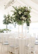 Elegant wedding reception with round tables, white chairs, and lush white floral centerpieces displayed on 32" tall rectangular gold metal flower stands with acrylic boards, all beneath a draped tent ceiling.