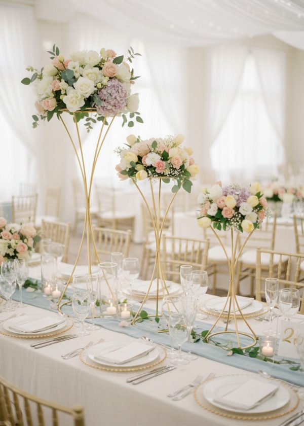 Elegant wedding table with floral centerpiece atop 40" gold hourglass pedestals (10 pack), surrounded by candles, glassware, white plates, and gold chairs in a bright room with flowing white drapery.