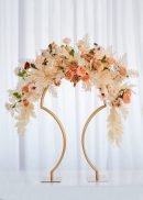 The 4 Pack Bridge Style Tabletop Centerpiece Matte Gold 31½" Tall holds peach and cream roses, white leaves, and greenery for a stunning display against a white curtain backdrop.