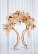 A stunning centerpiece is created using the 4 Pack Bridge Style Tabletop Centerpiece Matte Gold 31½" Tall, adorned with peach, cream, and blush flowers with greenery, set against a white curtain backdrop.