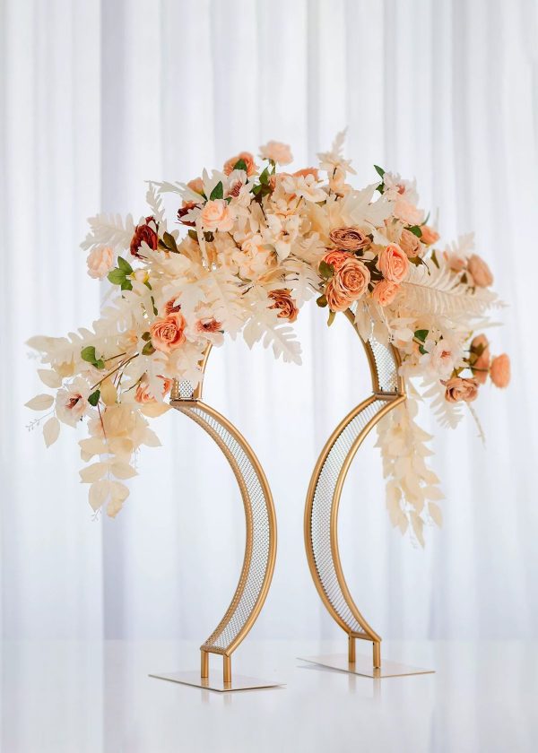 A stunning centerpiece is created using the 4 Pack Bridge Style Tabletop Centerpiece Matte Gold 31½" Tall, adorned with peach, cream, and blush flowers with greenery, set against a white curtain backdrop.