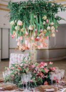 A lavish table centerpiece showcases the 2 Pack Floral Riser Tabletop Centerpiece, 4ft Tall in Gold, adorned with cascading tulips and pink roses among lush greenery, complemented by elegant glassware and refined table settings below.