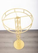 The 2 Pack Floral Riser Tabletop Centerpiece, each 4ft tall in gold, features a round base and open geometric frame made of intersecting bars, shown from above on a light wood floor against a white wall.