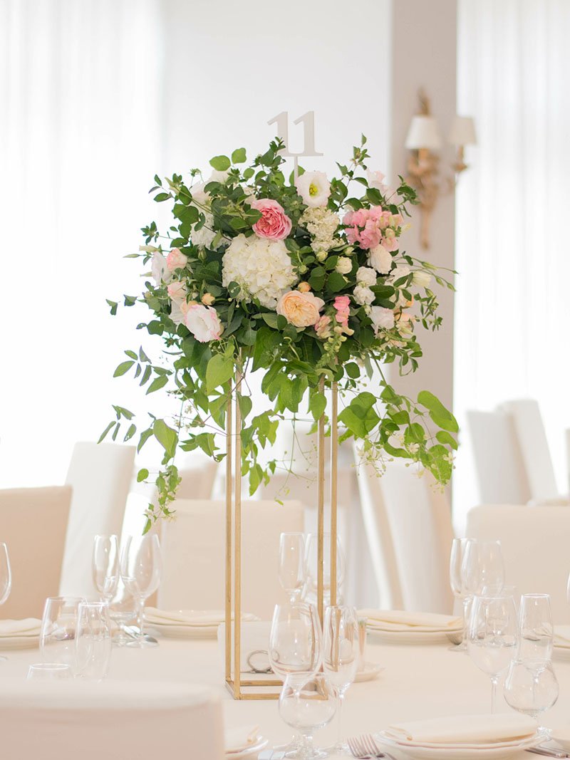 A tall floral centerpiece with pink, white, and green flowers sits on a gold stand in the center of a round table set with empty glassware and white napkins in a bright, elegant room. Table number 11 is displayed on top.