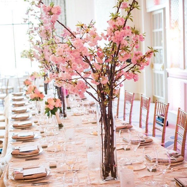 A long banquet table set for an event, decorated with tall centerpieces of pink cherry blossom branches, surrounded by wooden chairs, with elegant tableware and soft natural light filling the room.