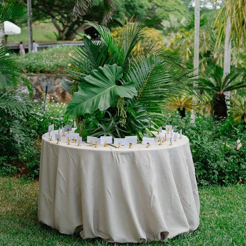 A round table with a beige tablecloth sits outdoors, topped with a large tropical leaf arrangement and surrounded by small white place cards held by gold holders. Lush greenery and trees are in the background.