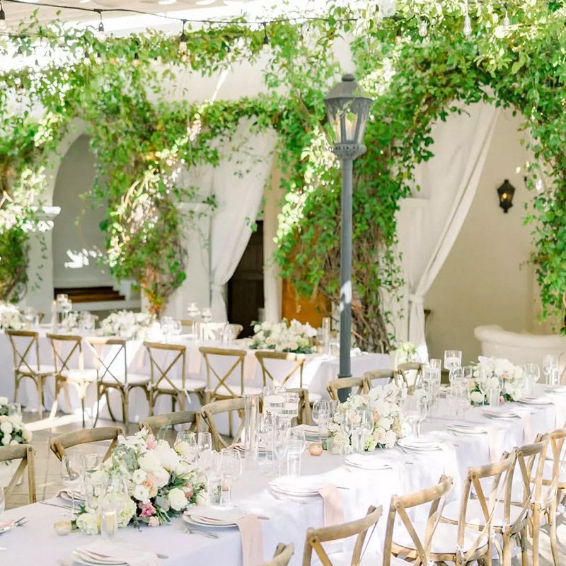 Elegant outdoor wedding reception with long tables covered in white tablecloths, floral centerpieces, and wooden chairs. Green vines and leaves drape over white arches, creating a romantic, garden-like atmosphere.