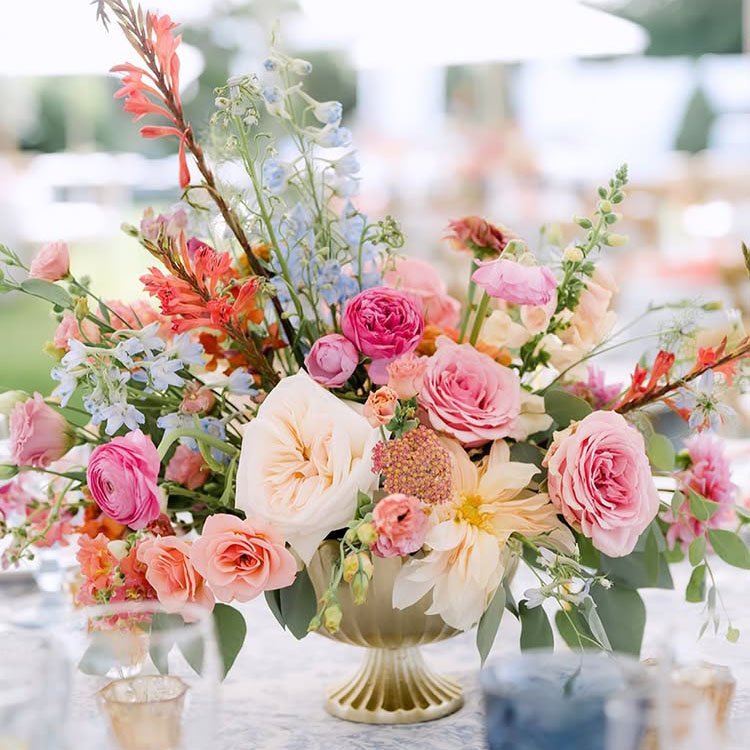 A decorative vase overflowing with a vibrant arrangement of pink, peach, and orange flowers, accented with green leaves, placed on a table outdoors.