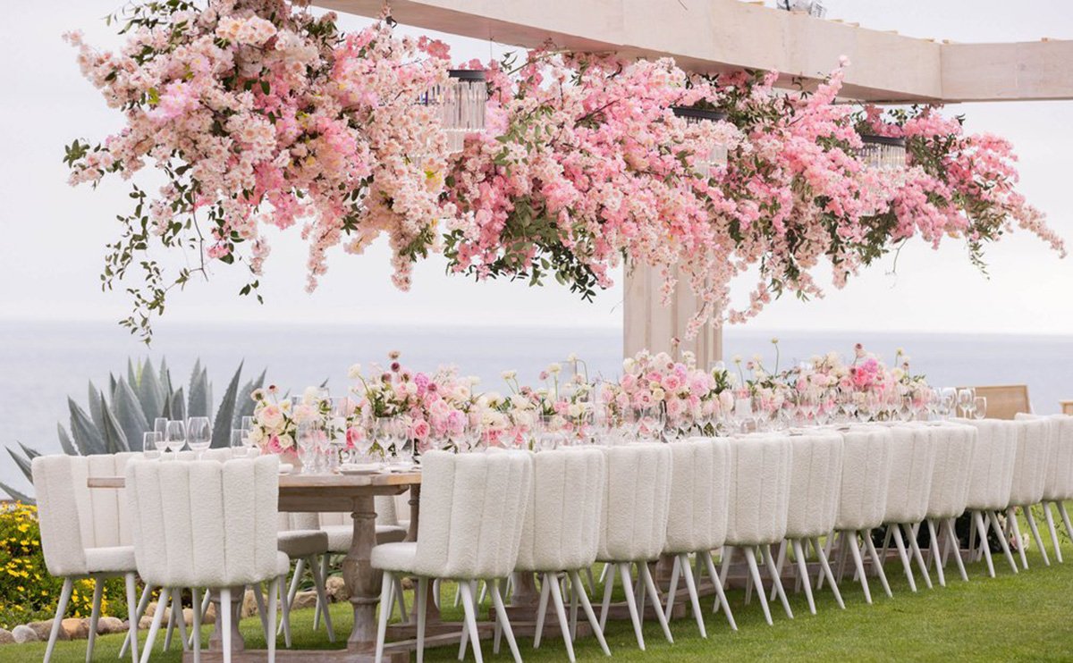 A long outdoor table with white cushioned chairs is elegantly set under an overhead arrangement of cascading pink flowers, with the ocean visible in the background.