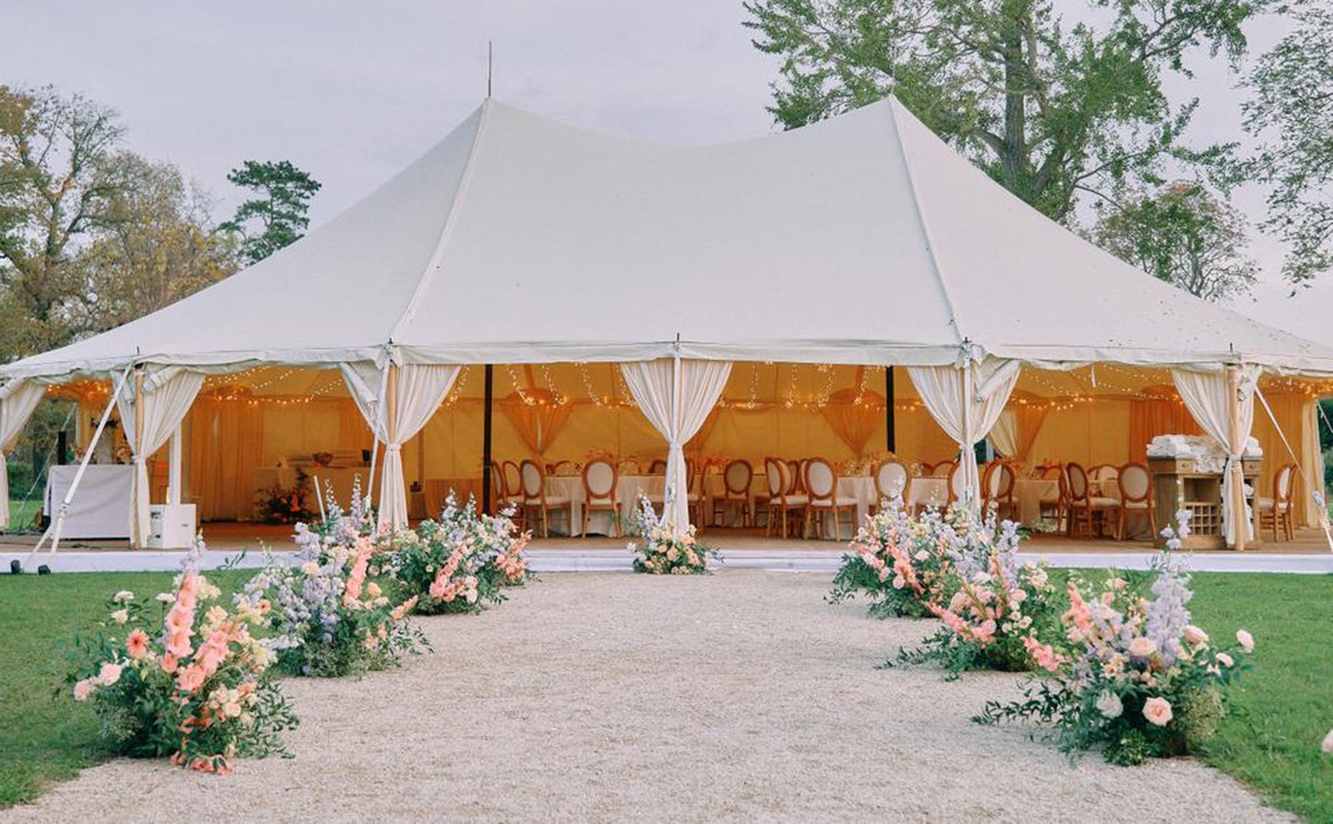A large white event tent with open sides stands on a lawn, decorated with string lights. Inside are elegant chairs and tables, and a display of UBIZ wedding gifts. A gravel path lined with colorful flower arrangements leads to the entrance.