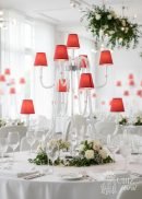 A bright, white room features a round wedding table set with the 44" tall, 13-arm Wedding Candelabra with red lampshades and LED lights, surrounded by white flowers, greenery, wine glasses, and cutlery.