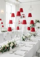 A modern dining table is elegantly set with white linens, glassware, floral arrangements, and the Wedding Candelabra 44" Tall—13-Arm with Red Lampshades LED Lights—centerpiece. The setting is bright and airy with natural light.