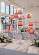A bright, elegant wedding table features the 44" tall 13-arm Wedding Candelabra with red lampshades and LED lights as a centerpiece, surrounded by white linens, floral arrangements, and formal place settings.