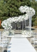 The Curved Pushup Backdrop Kit Wedding Backdrop Stage Centerpiece 17.9' Long features a white floral arch with cascading flowers and hanging ribbons, creating a stunning outdoor backdrop on a reflective platform surrounded by white chairs and greenery.