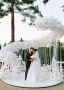 A bride and groom share an intimate moment at an outdoor wedding, standing under the Curved Pushup Backdrop Kit Wedding Backdrop Stage Centerpiece (17.9' long) adorned with white flowers and drapes amid lush greenery for a dreamy, romantic setting.