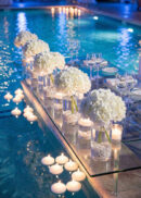 A glass table set with white hydrangea centerpieces, candles, and elegant place settings is positioned over a pool, surrounded by floating candles, creating a romantic and luxurious atmosphere.