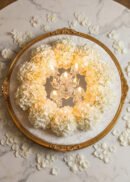 A small chandelier surrounded by a circular wreath of white hydrangeas sits on a marble table, with scattered white petals around the arrangement and a decorative gold frame encircling the flowers.