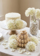 A white-frosted cake on a stand, a pyramid of beige macarons, and glass vases with white hydrangeas and white candy sit on a table decorated with white flower petals and hydrangea blooms.