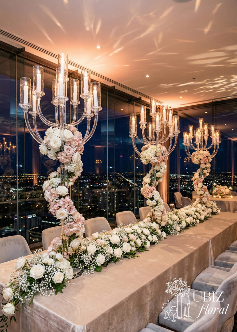 A 13-Head Clear Crystal Candelabra Wedding Table Centerpiece, adorned with white and blush roses, stands on a banquet table lined with matching floral garlands in a modern glass-walled venue overlooking the sparkling city at night.