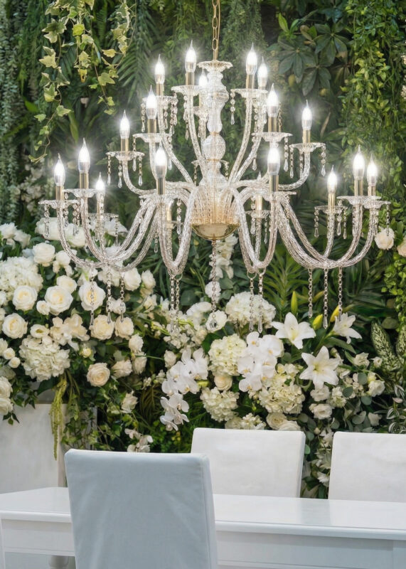 A 2.5ft Silver 20-Light 3-Tier Crystal Chandelier with classic curved arms and crystal drops hangs above a white dining table and chairs, set against lush green foliage and white flowers—perfect for weddings and events.