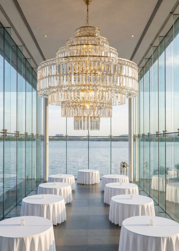 Elegant banquet hall with floor-to-ceiling glass walls overlooking the water, featuring round tables with white linens and a 2.7ft Gold 7-Tier Round Crystal Chandelier with Prism Rods illuminating the space.