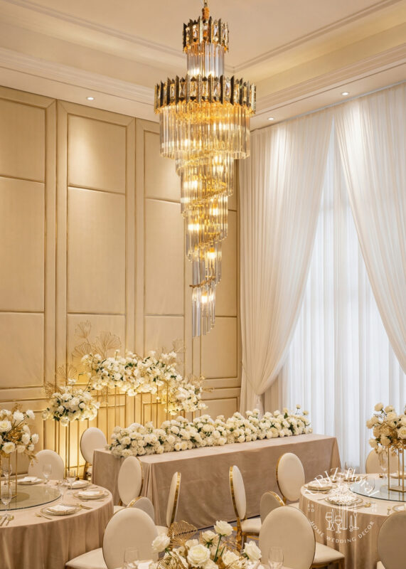 An elegant wedding reception features cream and gold decor, white floral centerpieces, and a 5.8’ tall x 2.4’ wide Gold Spiral Crystal Rod Chandelier glowing above the head table. Tall windows with sheer curtains complete the sophisticated look.