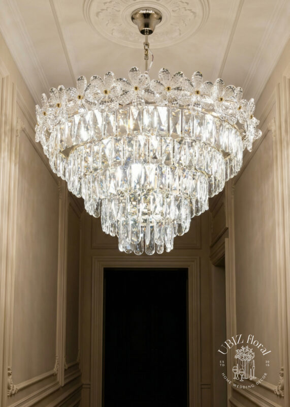 The 12"H x 16"W Crystal Daisy Crown Tiered Chandelier in Silver & Gold decorates an elegant hallway with ornate molding. Multiple tiers of hanging glass prisms shimmer, and the UBLZ Floral logo appears in the bottom right corner.