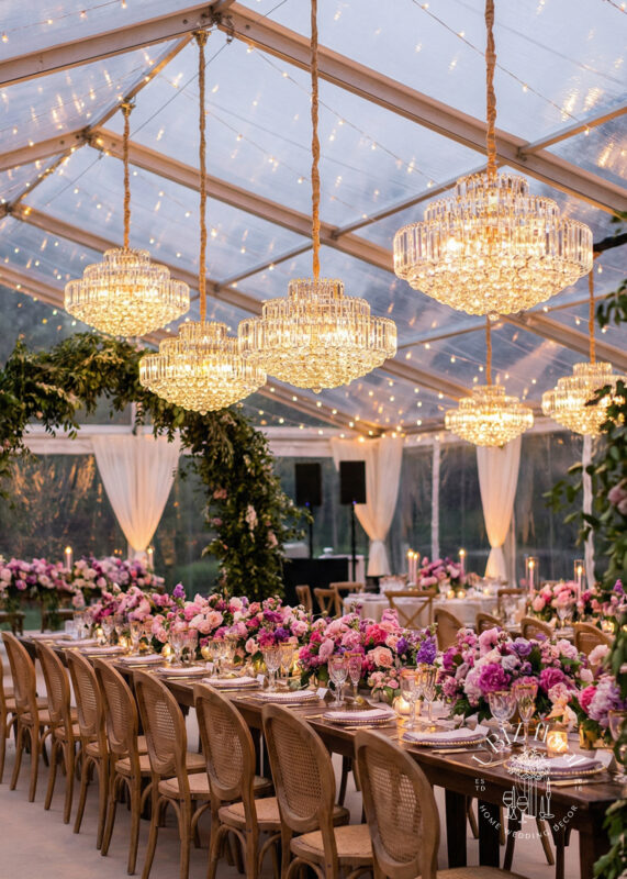 A luxurious wedding reception under a clear tent, adorned with 24” Gold Tiered Crystal Drum Chandeliers with Crystal Sphere Bases, wooden chairs and long tables, lavish pink and purple floral centerpieces, and romantic string lights overhead.