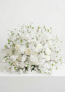 A large, elegant floral centerpiece featuring white roses and delicate pink blossoms with green leaves, displayed against a plain white background.