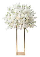 A large white flower arrangement, featuring roses and blossoms, forms an elegant centerpiece atop a tall gold rectangular stand, with hints of pink accents displayed against a white background.