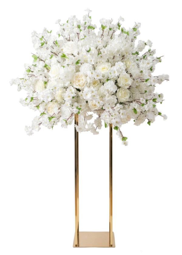 A large white flower arrangement, featuring roses and blossoms, forms an elegant centerpiece atop a tall gold rectangular stand, with hints of pink accents displayed against a white background.