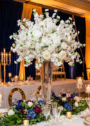 A tall, elegant vase serves as a stunning centerpiece, holding a large arrangement of white flowers and roses, surrounded by candles and smaller floral decorations with purple and pink accents on a lavishly set banquet table.