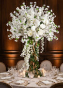 A tall floral arrangement with white roses, cherry blossoms, and touches of pink stands on a round table set with white dishes, crystal glasses, napkins, and surrounded by lit candles; the background features dark wood paneling.