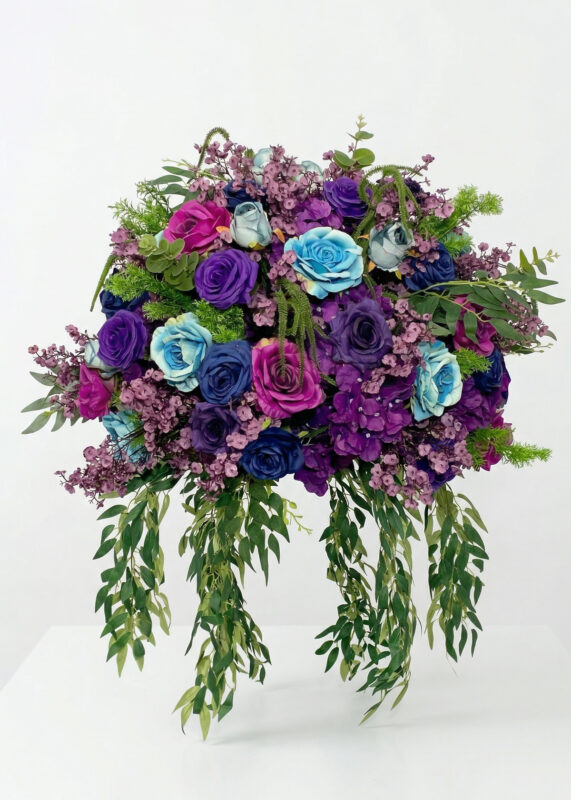 A vibrant floral centerpiece featuring blue, purple, and magenta roses, accented with green leaves and trailing greenery, displayed against a plain white background.
