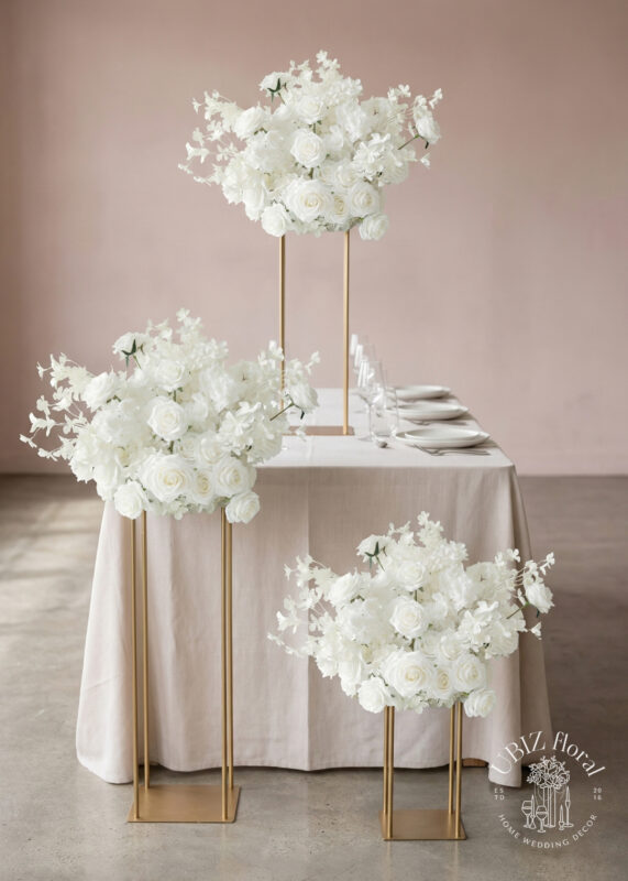 Three elegant gold stands hold lush flower ball centerpieces of white blooms, including roses and orchids, beside a rectangular table set for six, against a soft pink wall. A logo for UBIZ floral decor appears in the lower right corner.