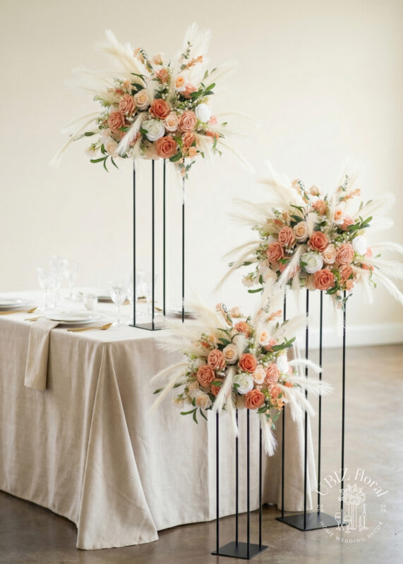 Elegant wedding table with beige tablecloth, set for guests, featuring a stunning wedding centerpiece: three tall black stands holding large arrangements of peach and white roses, white pampas grass, and greenery.