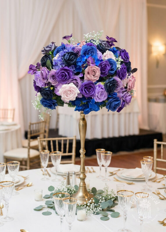 A tall gold centerpiece holds an arrangement of purple, blue, blush, and pink roses on a round table set with elegant glassware, gold-rimmed plates, and greenery in a decorated event venue with draped white curtains.