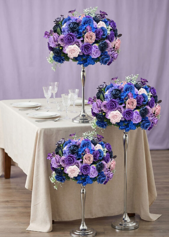 Three tall silver centerpiece stands hold spherical arrangements of purple, blue, pink, and blush roses with white babys breath. A table set with glassware and plates is covered with a beige cloth, and a lavender backdrop is behind.
