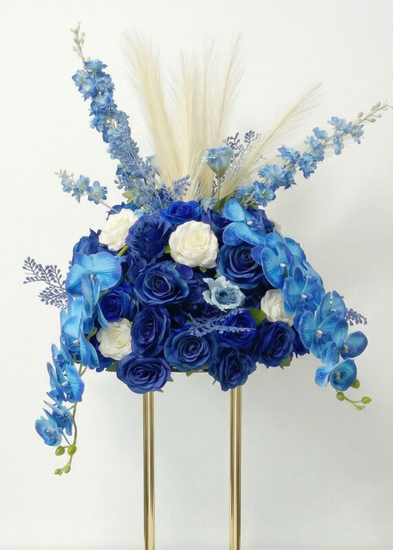 A stunning flower ball centerpiece featuring blue and white roses, blue orchids, delicate white pampas grass, and a hint of pink, all displayed on a gold stand against a plain white background.