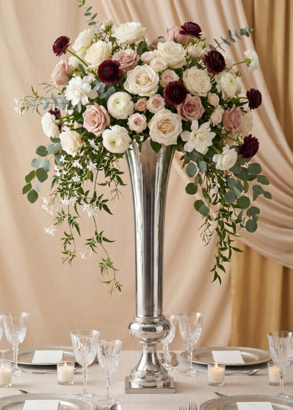 A 40in Silver Metal Trumpet Column Flower Vase Radiant Elegant for Wedding Event Flower Ball Centerpieces, filled with white, blush, and burgundy roses and eucalyptus, stands on a table set with candles, glassware, plates, menus. Draped fabric forms the backdrop.