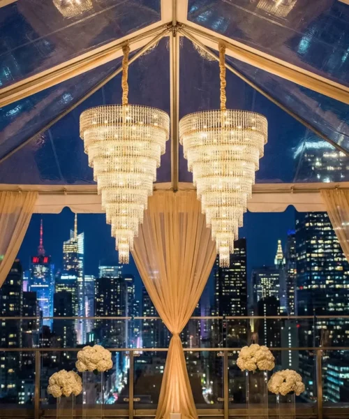 Elegant wedding venue with two large crystal chandeliers, sheer gold drapes, white floral arrangements, and a clear tent ceiling overlooking a city skyline at night.