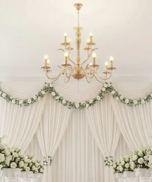 Elegant wedding ceremony setup with white draped curtains, green and white floral garlands, flower arrangements on white pillars, and a gold chandelier hanging from the ceiling.