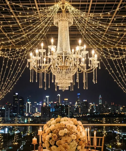 A crystal chandelier and string lights hang above a table with a large floral centerpiece and candles, set on a rooftop with a city skyline glowing at night in the background.