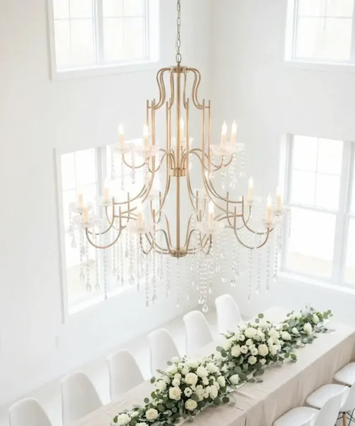 A long dining table with a beige tablecloth is decorated with white flowers and greenery, surrounded by white chairs, beneath a large, elegant chandelier in a bright, airy room with tall windows.