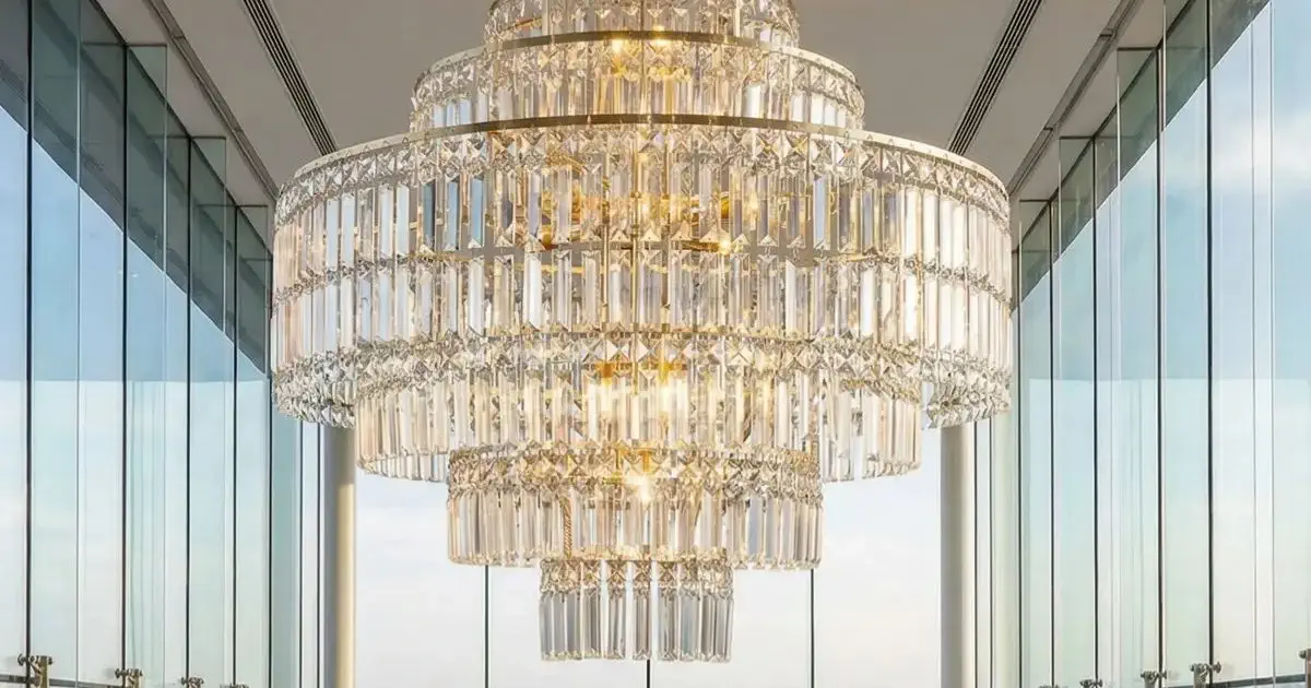 A large, elegant crystal chandelier hangs from a high ceiling between tall glass windows, reflecting natural light and creating a luxurious atmosphere.