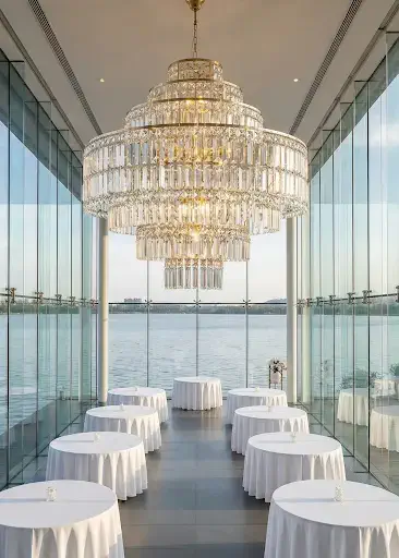 A large, elegant crystal chandelier hangs in a glass-walled room overlooking water. Several round tables with white tablecloths are evenly spaced beneath the chandelier.