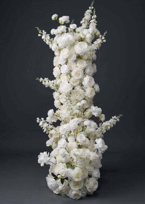 A tall, elaborate floral arrangement featuring layers of white roses and other white flowers, perfect as a wedding flower runner. The densely clustered blooms create a lush and elegant display against a dark gray background.