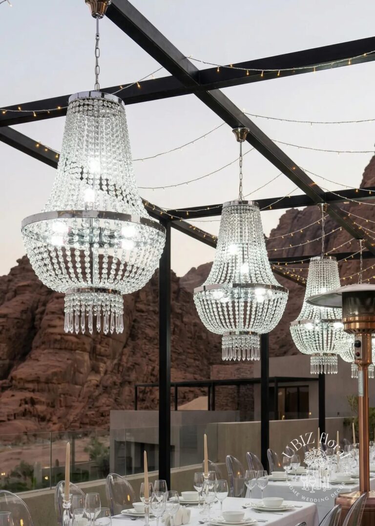 Elegant crystal chandeliers hang from a pergola decorated with string lights, set against a rocky mountain backdrop above an outdoor dining table set for an event.